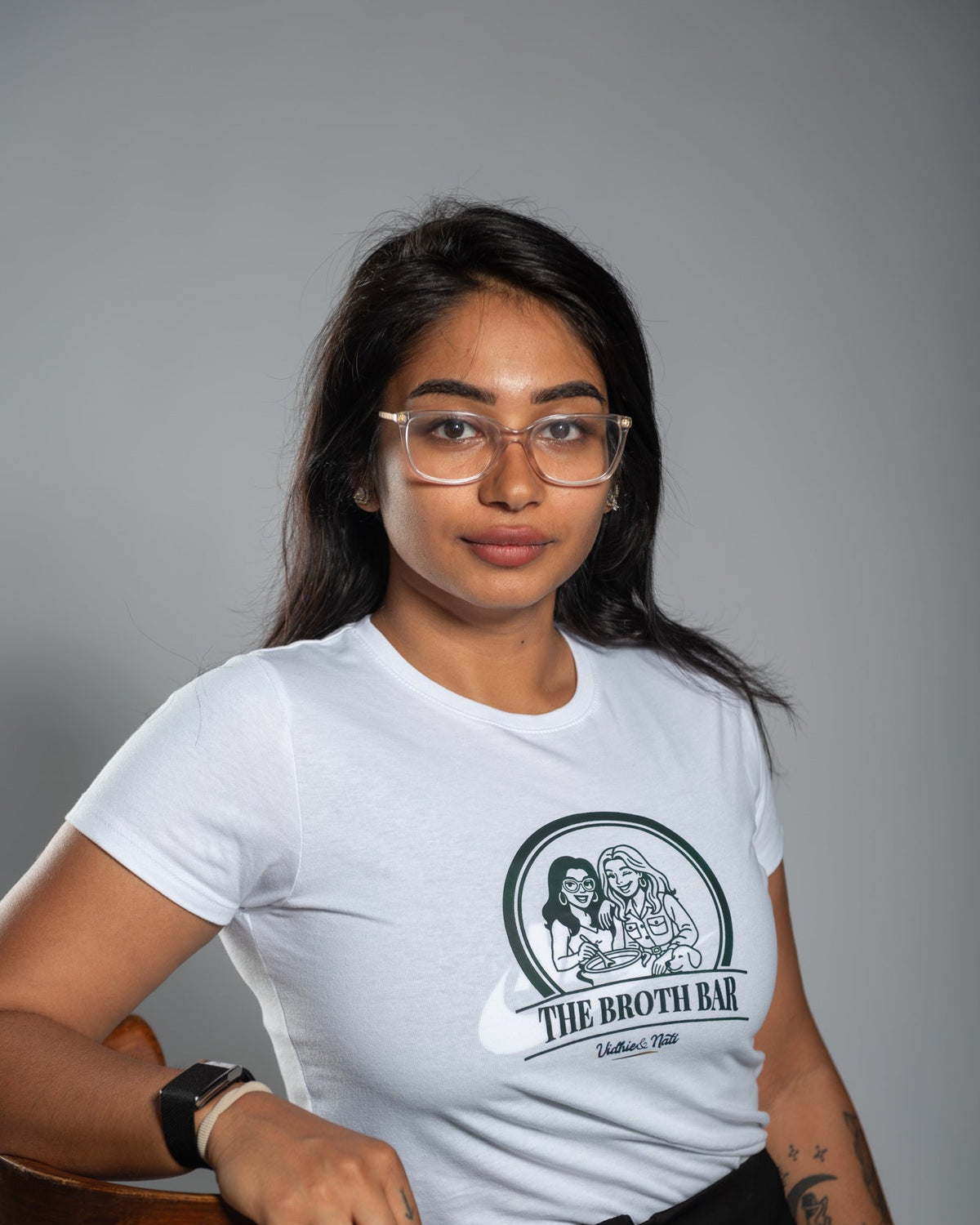 Woman wearing a white t-shirt with a logo and text, sitting on a wooden stool against a gray background.